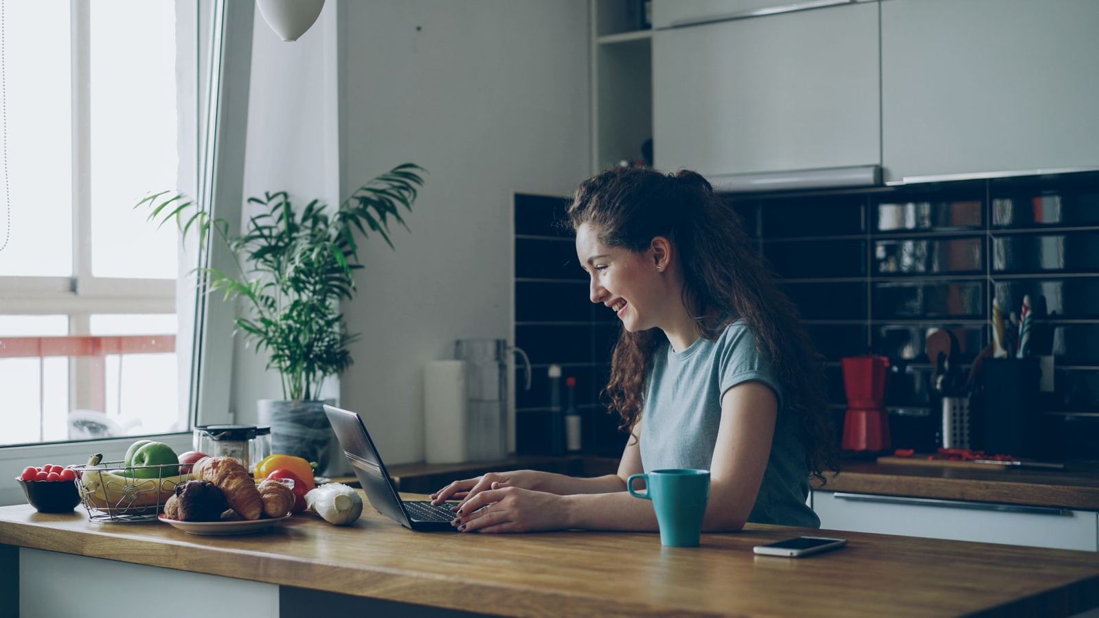 A woman laughing while working on her laptop at a kitchen table.